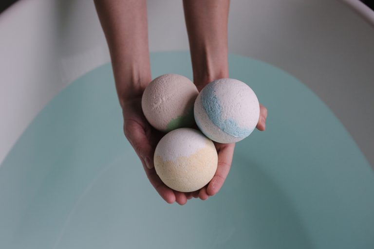 A pair of hands gently holding three natural-colored, handmade bath bombs (one cream/green, one white/blue, and one white/yellow) directly over a clean white bathtub filled with calm, light blue water.